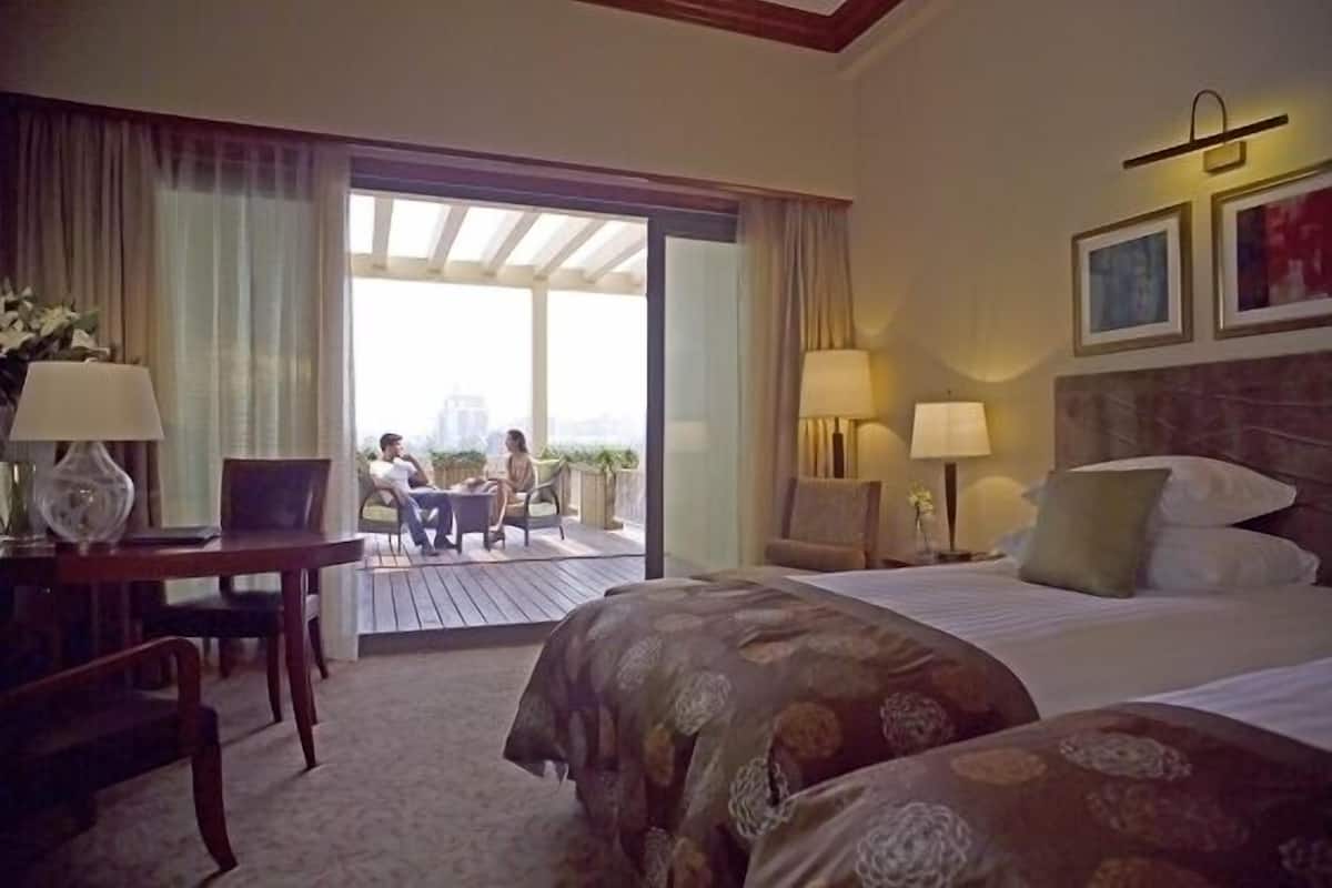Habitación de hotel con cama, dos mesitas de noche con lámparas, una silla, un escritorio y un balcón con vistas.