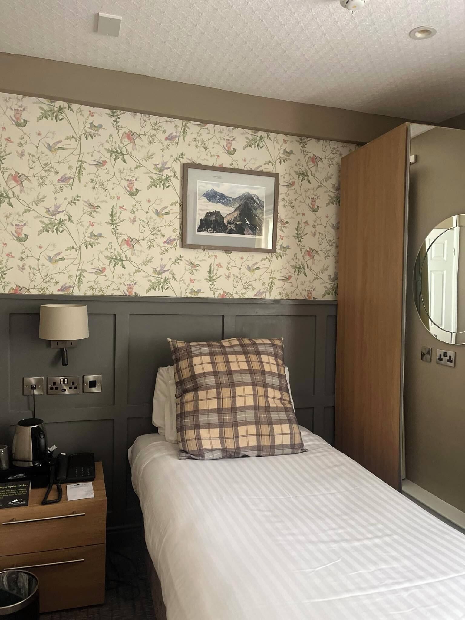 A hotel room with a bed, a nightstand, a lamp, a framed picture on the wall, and a wardrobe.