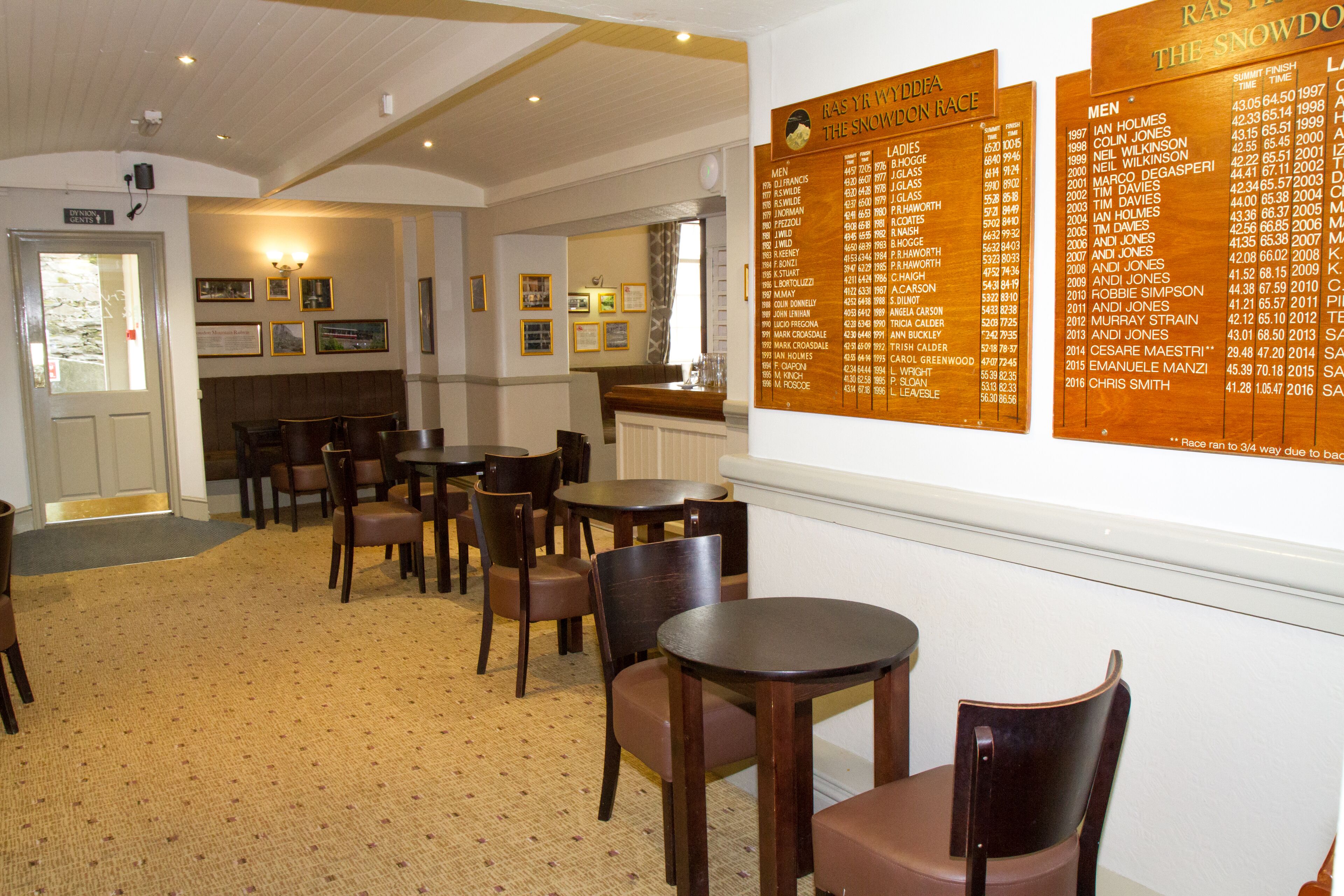 A dining area with wooden tables and chairs, a menu board on the wall, and framed pictures on the wall.