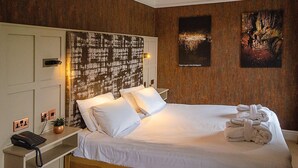 A neatly made bed with white linens, a wooden headboard, and a wall-mounted artwork depicting a cave scene. - The Royal Victoria Hotel (Caernarfon)