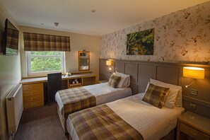 A hotel room with two beds, a desk, a chair, a radiator, a window, and a painting on the wall. - The Royal Victoria Hotel (Caernarfon)