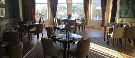 A dining area with multiple tables and chairs, large windows with curtains, and a view of the outdoors.