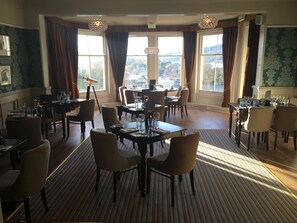 A dining area with multiple tables and chairs, large windows with curtains, and a view of the outdoors. - The Royal Victoria Hotel (Caernarfon)