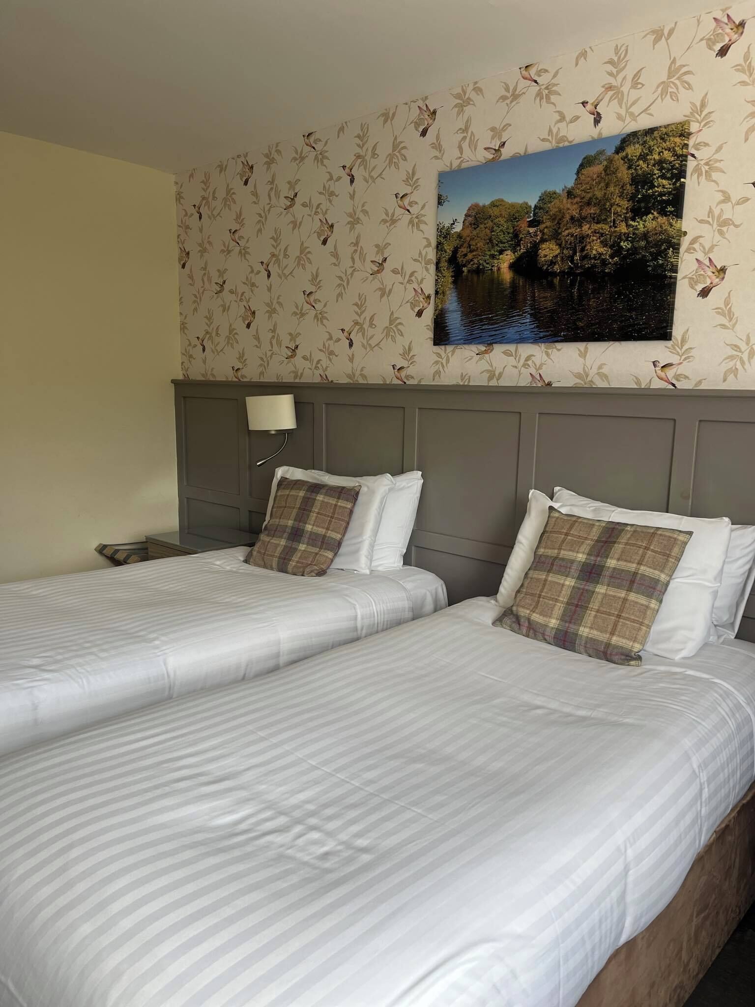 A hotel room with two beds, a nightstand, a lamp, and a wall-mounted painting of a landscape.