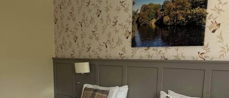 A hotel room with two beds, a nightstand, a lamp, and a wall-mounted painting of a landscape.