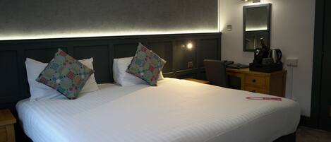A hotel room with a large bed, two pillows, a bedside table, a chair, a desk, a mirror, and a nightstand.