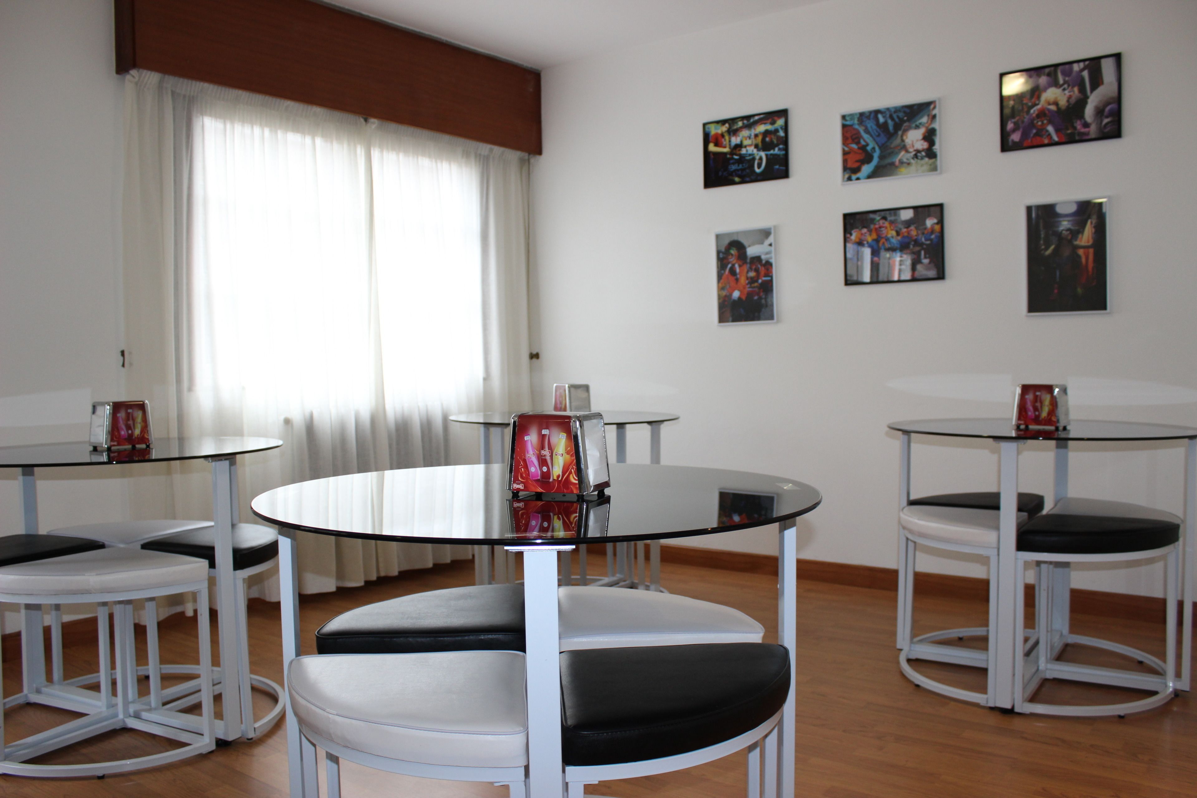 Una habitación con cuatro mesas redondas, cada una con tapa de cristal y estructura metálica blanca, rodeadas de sillas blancas con cojines negros. En la pared hay cuatro cuadros enmarcados, y se ve una ventana con cortinas blancas.
