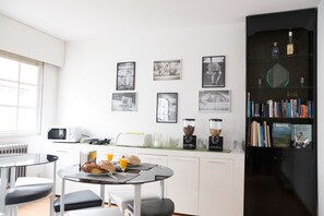 A modern kitchen with a dining area, featuring a coffee machine, toaster, and a bookshelf with books and decorative items.