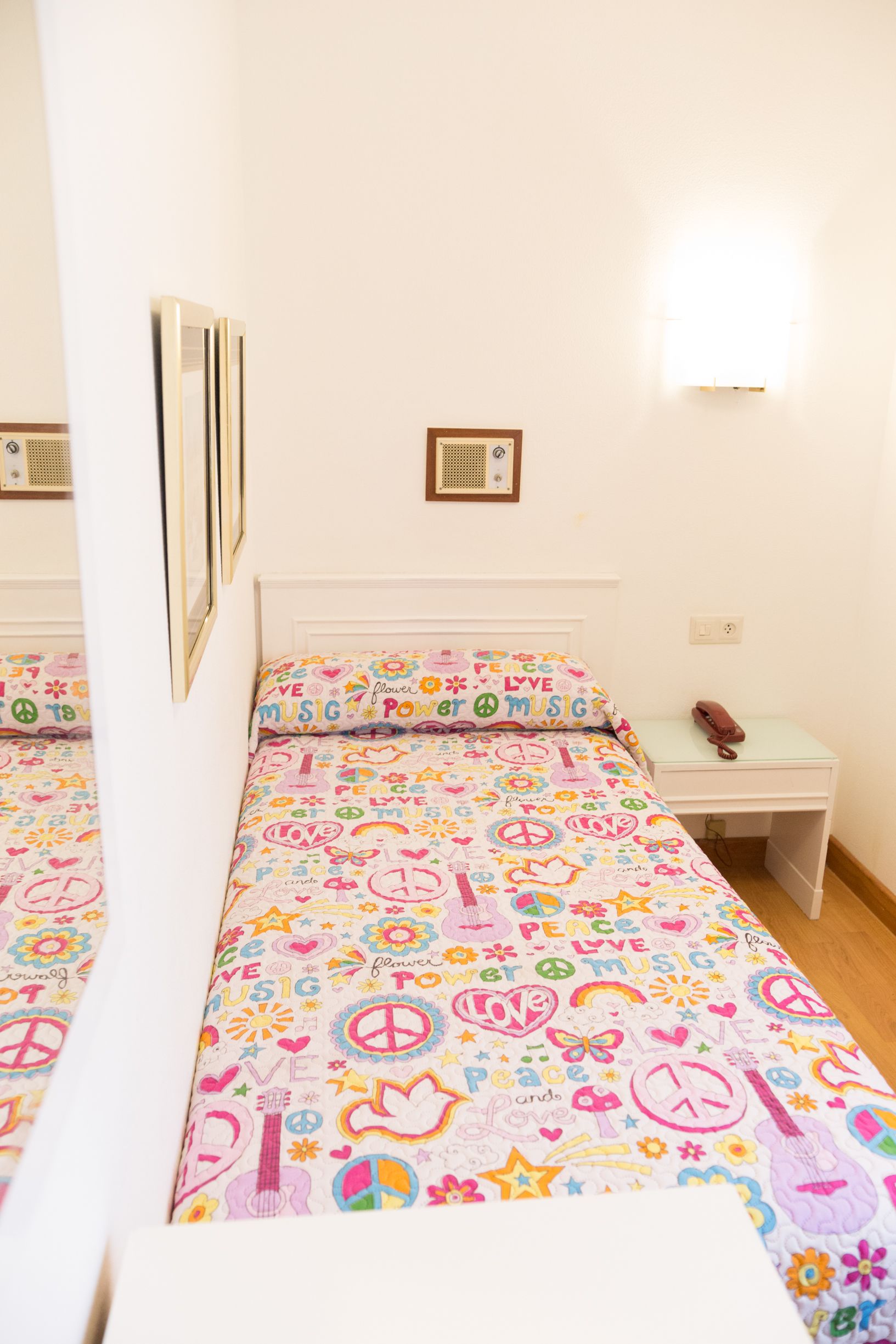 A bedroom with a bed covered in a colorful, patterned bedspread, a bedside table with a lamp, and a wall-mounted light fixture.