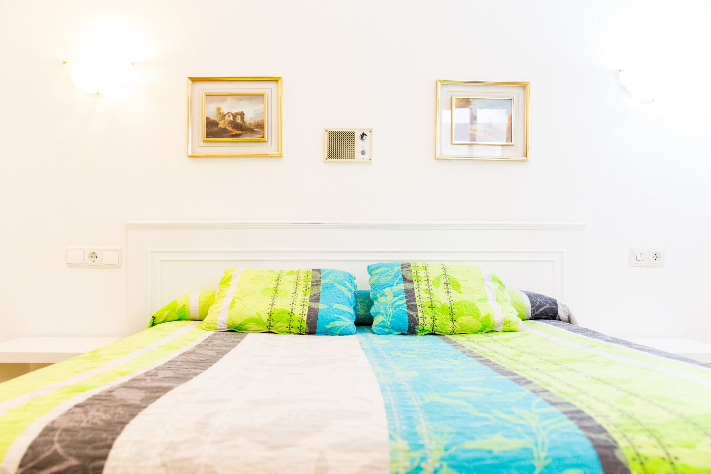 A bedroom with a bed, two framed pictures on the wall, and a power outlet.