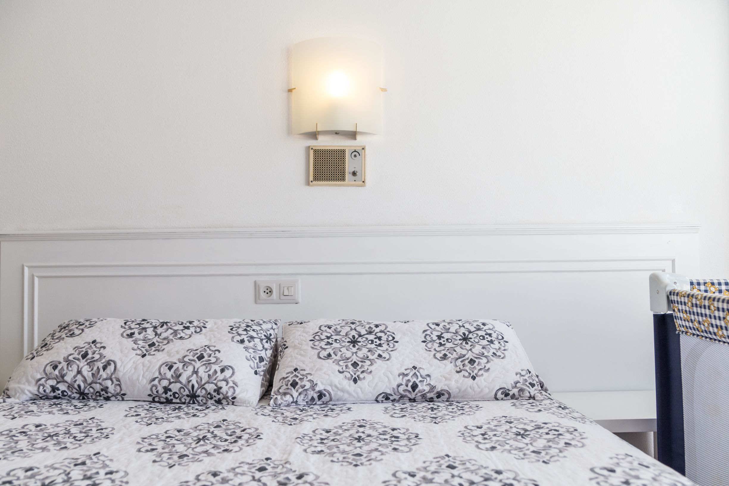 A neatly made bed with patterned bedding, a wall-mounted light fixture, and a small ventilation unit.