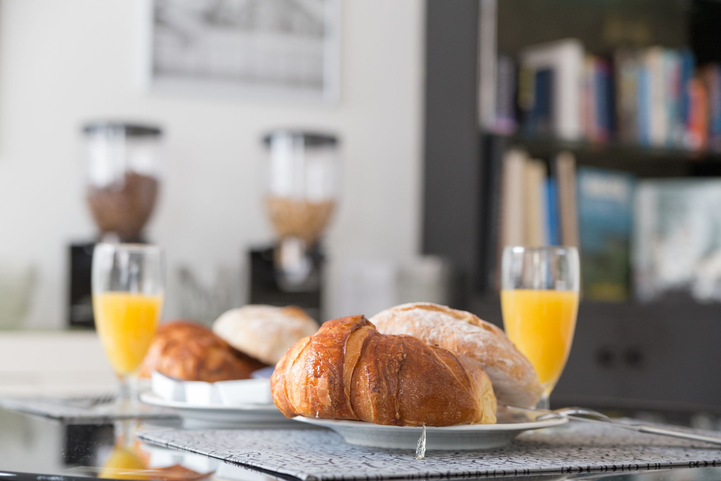 Croissants on a plate with a glass of orange juice, a coffee maker, and a bookshelf in the background.