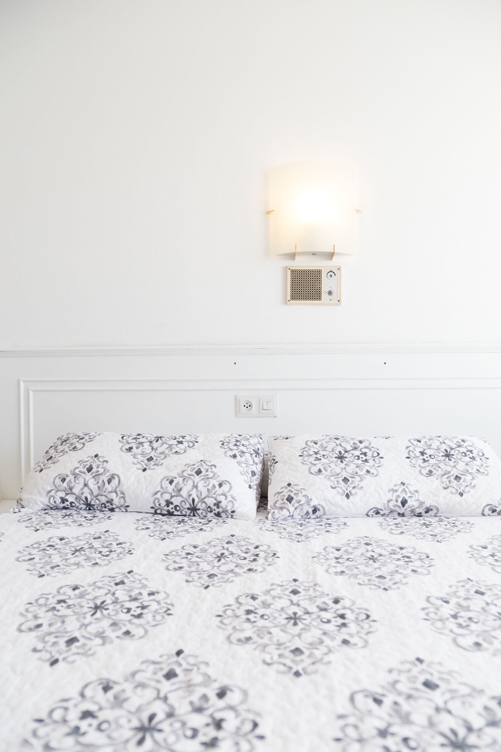 Una cama con colcha estampada, cabecero blanco y lámpara de pared.