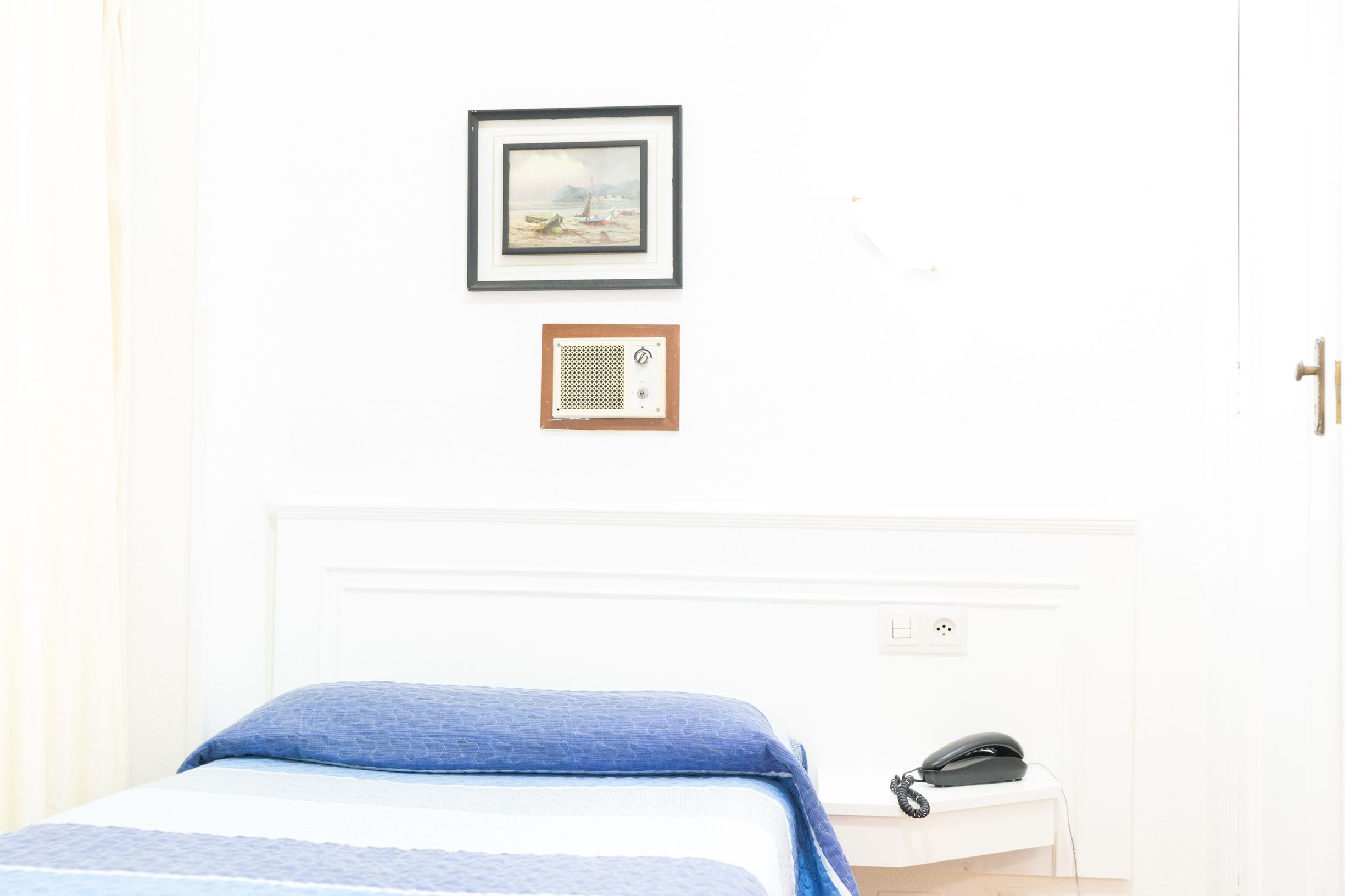A bedroom with a bed, a bedside telephone, a framed picture, and a wall socket.