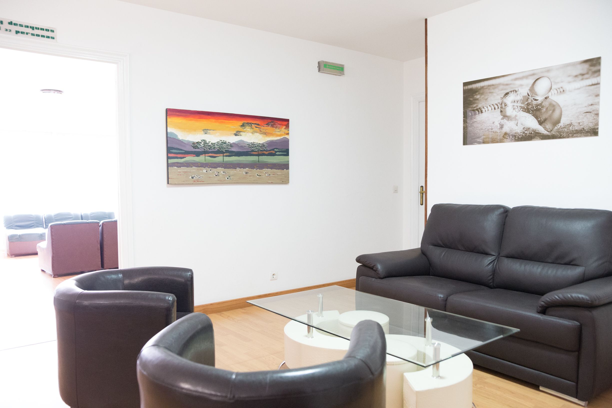 A modern living room with a leather sofa, a glass coffee table, and two armchairs. There are two paintings on the walls, one depicting a landscape and the other a swimmer.