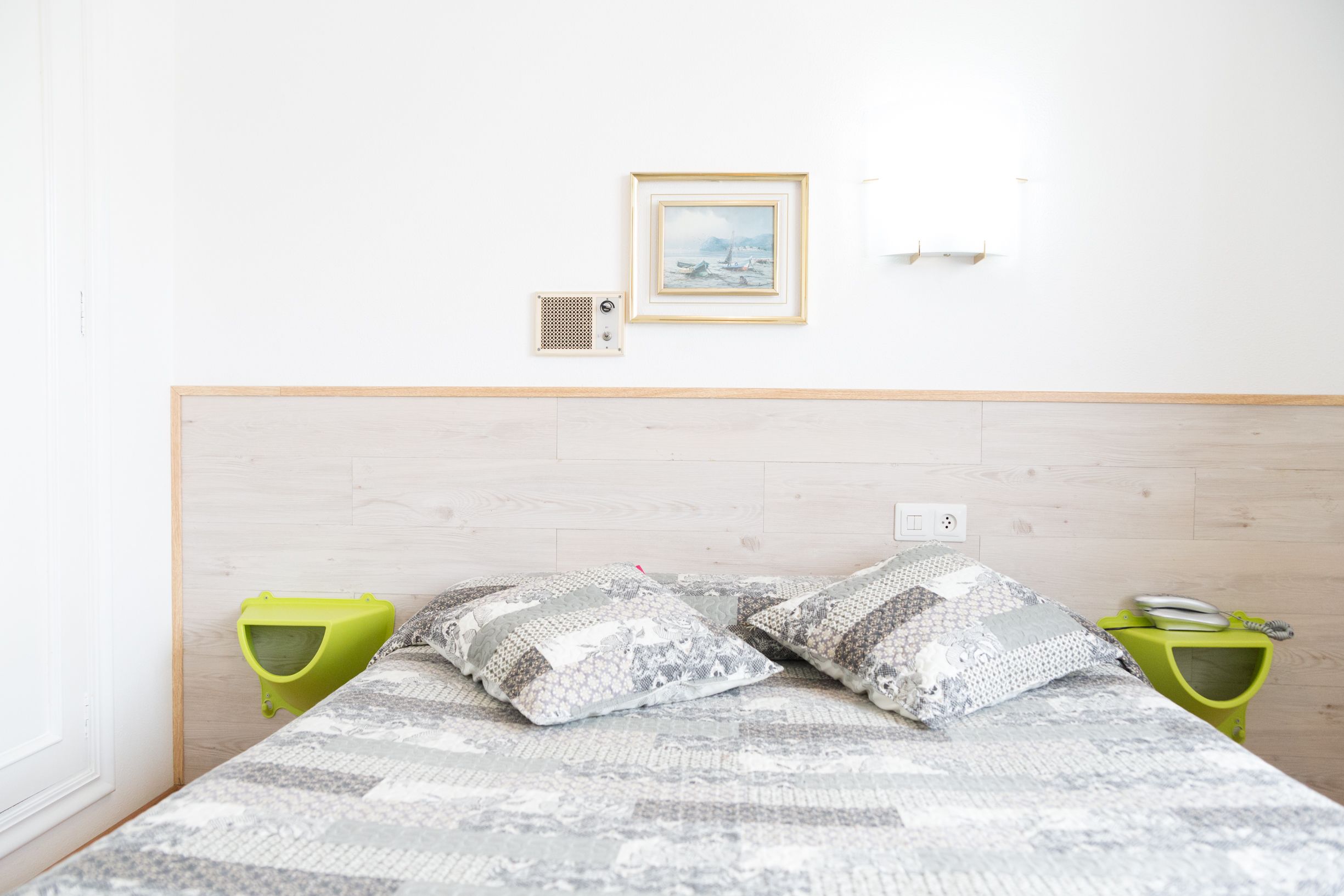 Un dormitorio ordenado con cama, almohadas, un cuadro enmarco, un calefactor en la pared y una mesita de noche verde con una lámpara.