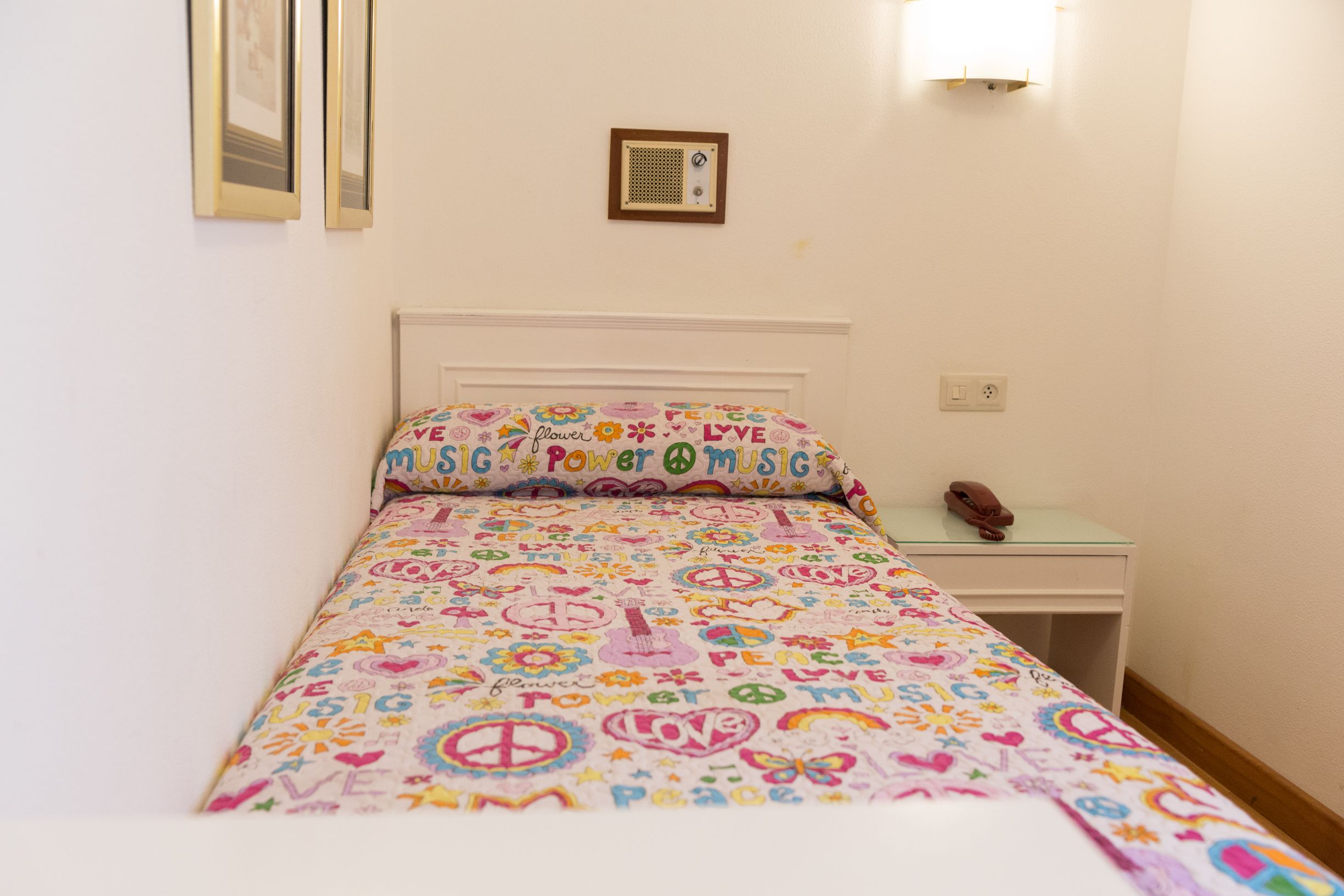 A bedroom with a bed covered in a colorful blanket featuring peace symbols and the words "LOVE" and "MUSIC". There is a small bedside table with a lamp and a phone.
