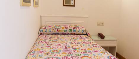 A bedroom with a bed covered in a colorful blanket featuring peace symbols and the words "LOVE" and "MUSIC". There is a small bedside table with a lamp and a phone.