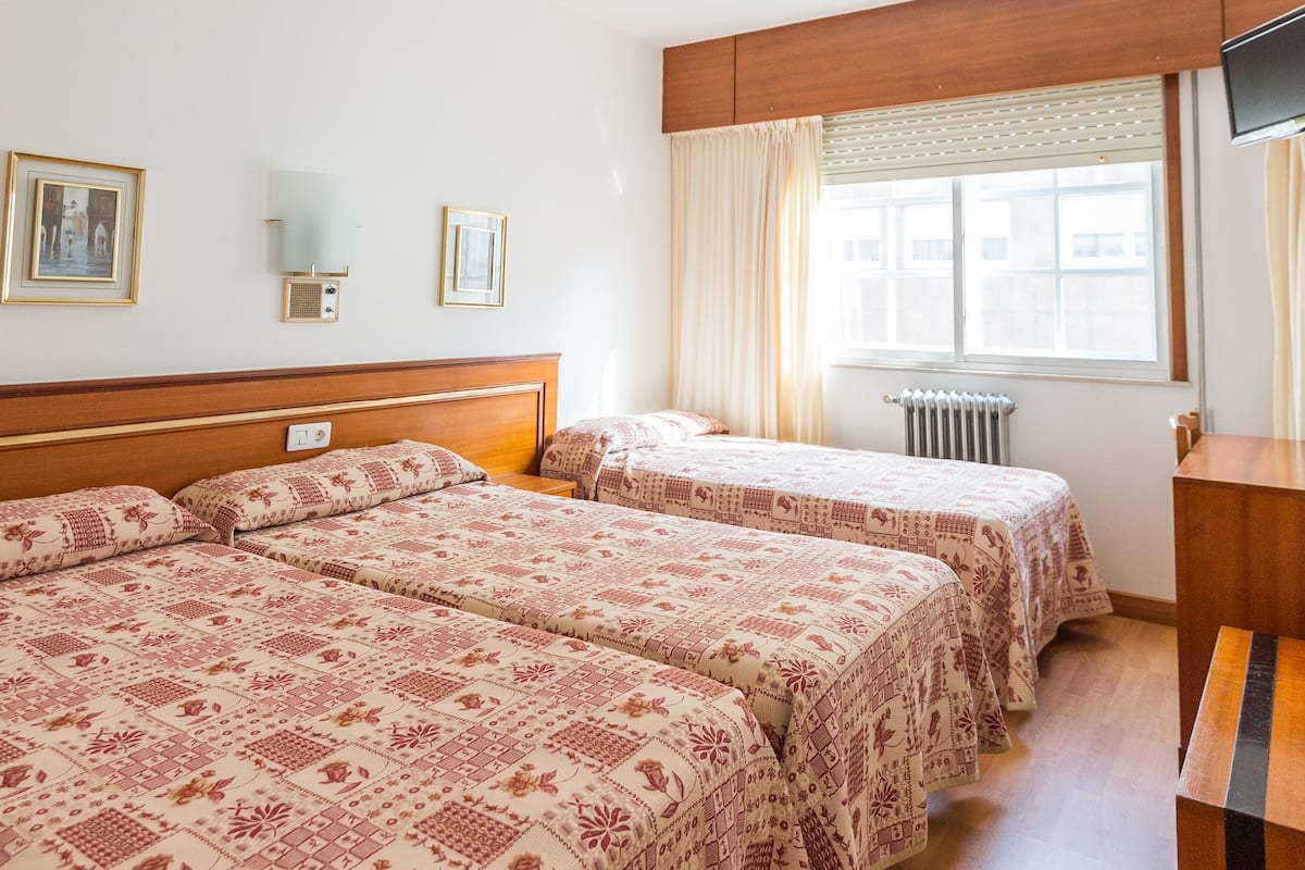 A hotel room with two beds, a wooden headboard, a window with curtains, a radiator, and a flat-screen TV mounted on the wall.