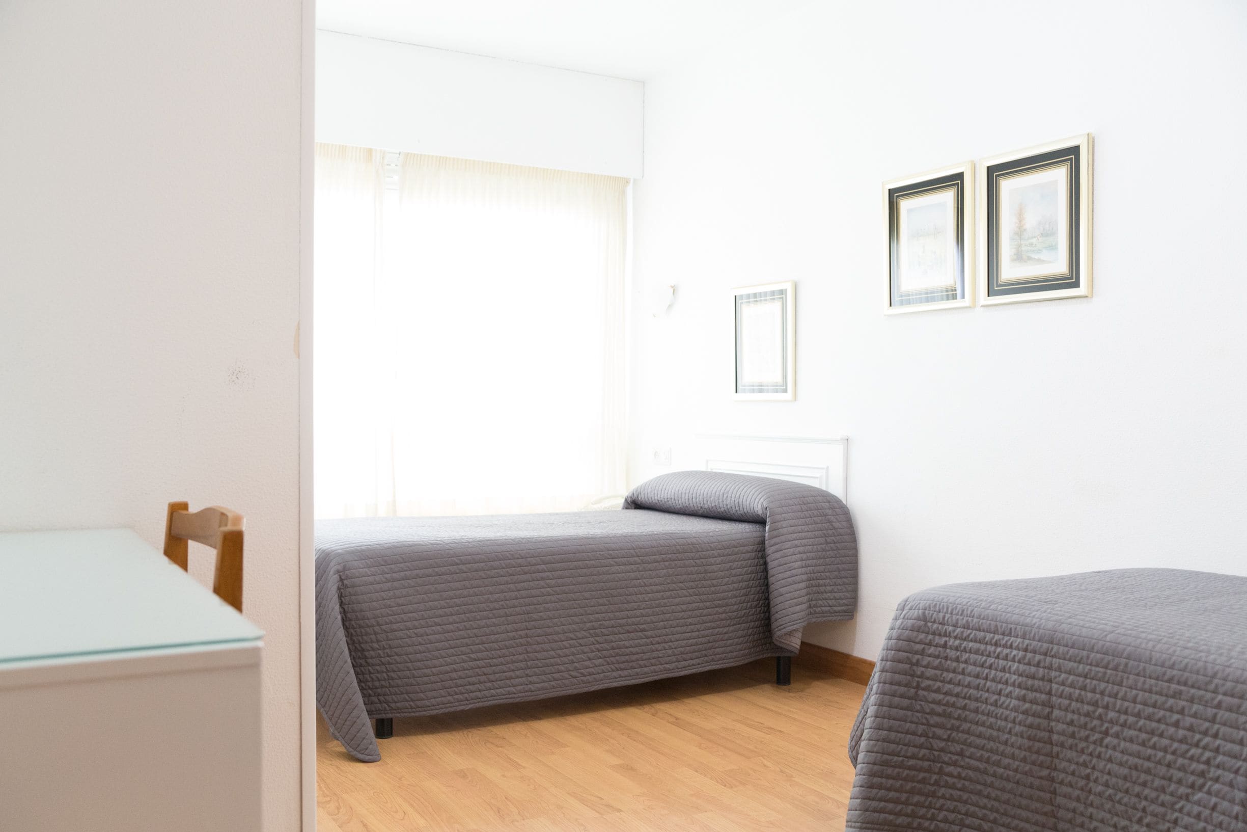 Una habitación pequeña y luminosa con dos camas, una mesa de madera y un ventanal con cortinas de gasa.