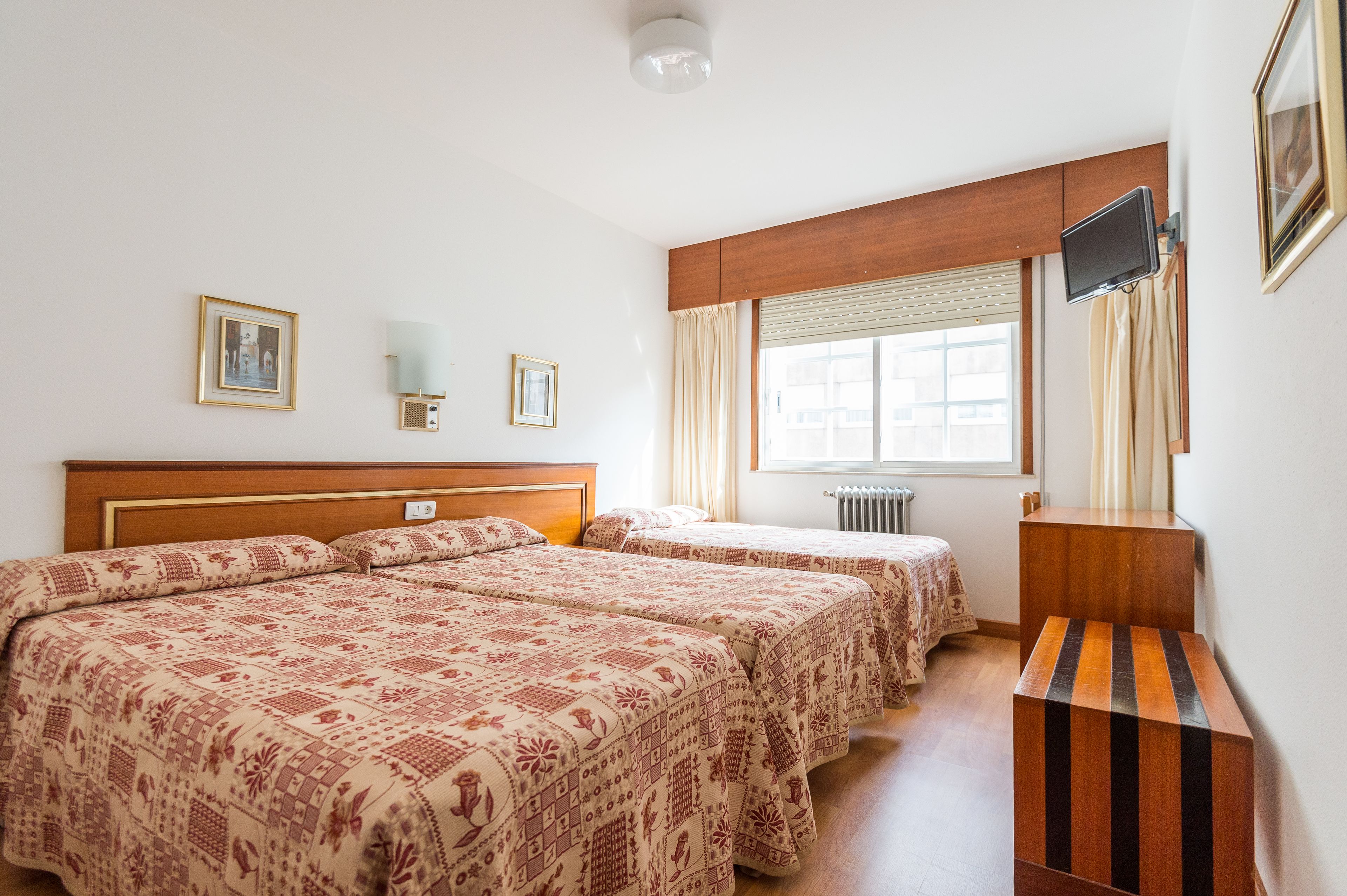 A hotel room with two beds, a wooden headboard, a TV mounted on the wall, and a window with curtains.