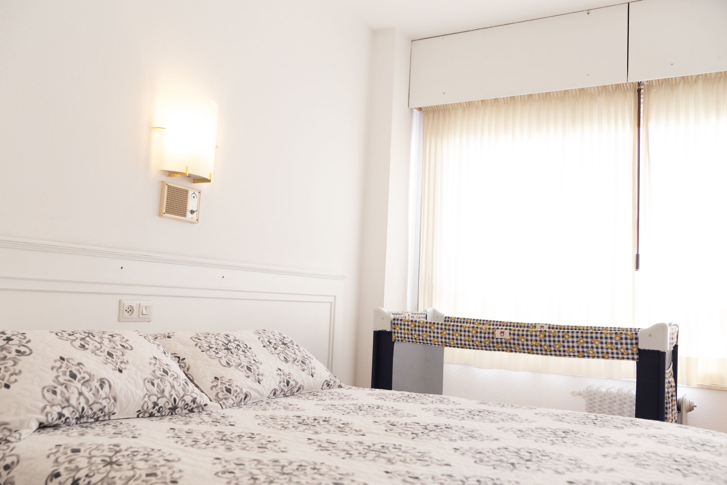 A hotel room with a bed, a wall-mounted light, a window with curtains, and a small table with a chair.