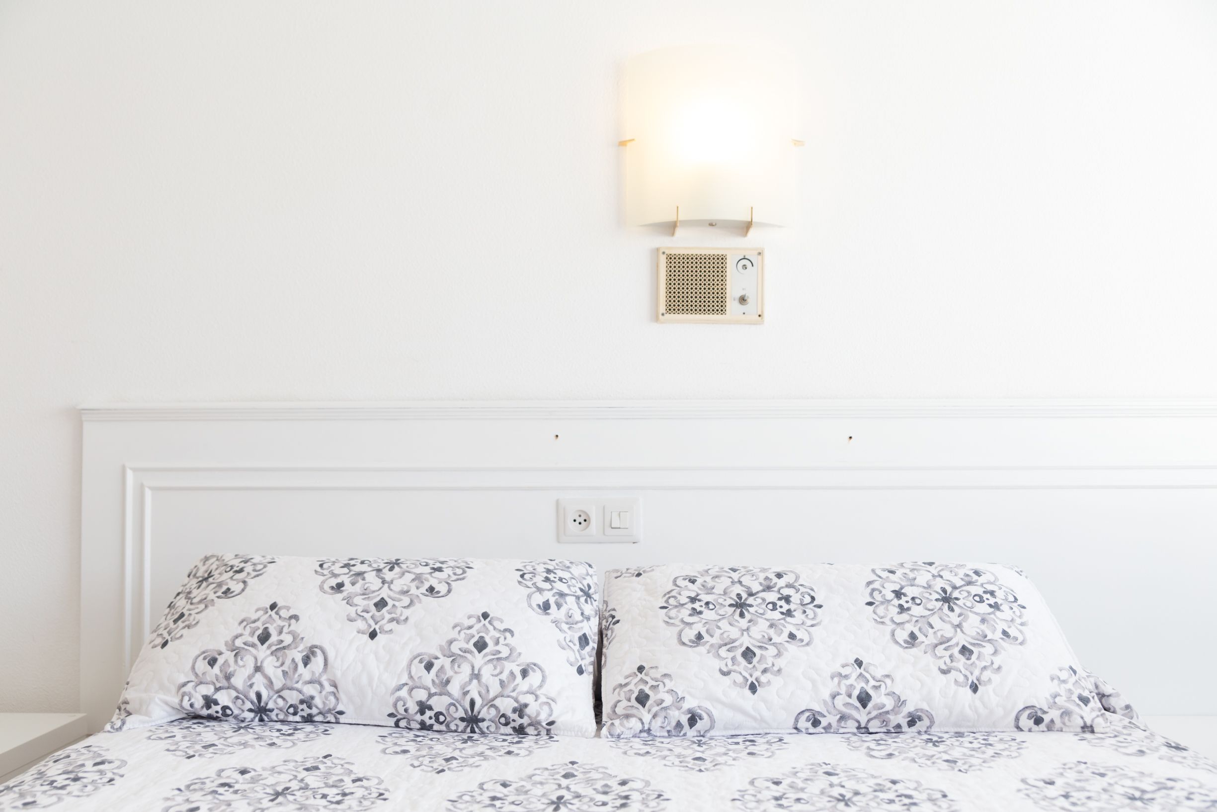 Una cama bien tendida con ropa de cama estampada, una lámpara de pared y un pequeño altavoz.