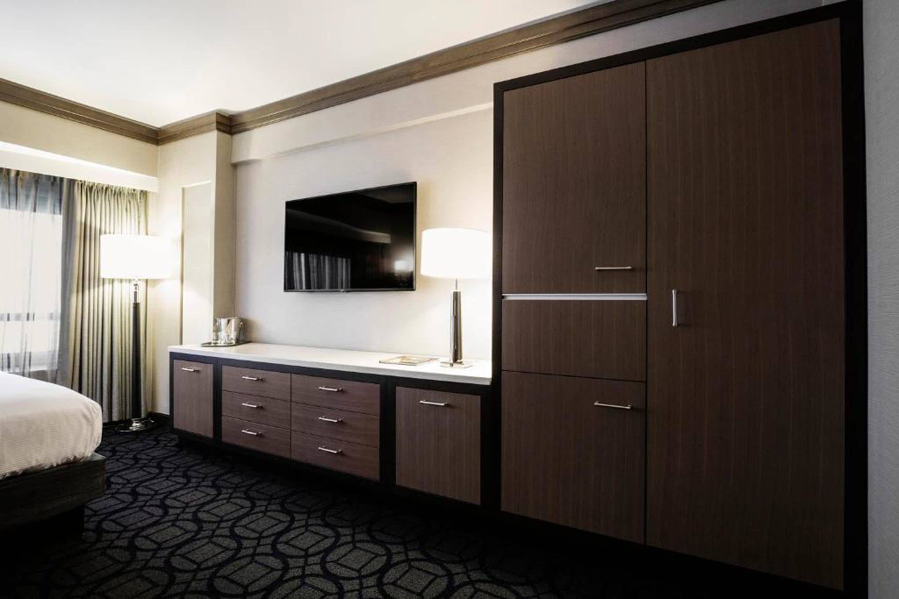 Habitación de hotel con cama, escritorio con cajones, armario grande, televisor de pantalla plana montado en la pared y alfombra estampada.