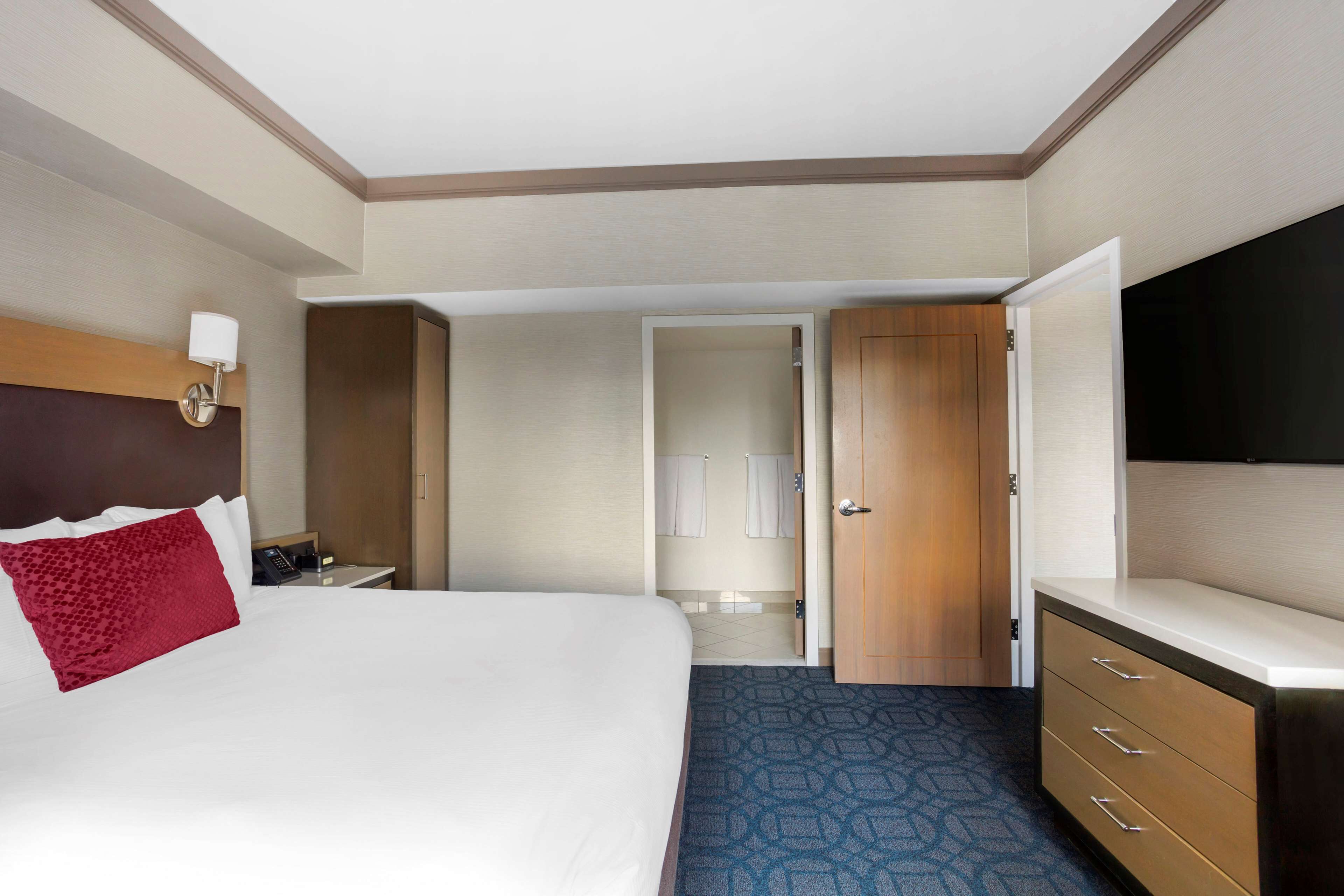 A hotel room with a large bed, a wooden headboard, a bedside table with a lamp, a dresser, and a flat-screen TV mounted on the wall.