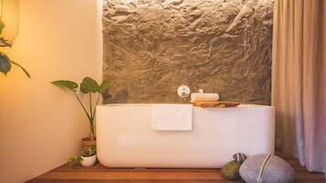 A bathroom with a stone wall, a white bathtub, a towel, a potted plant, and a wooden floor.