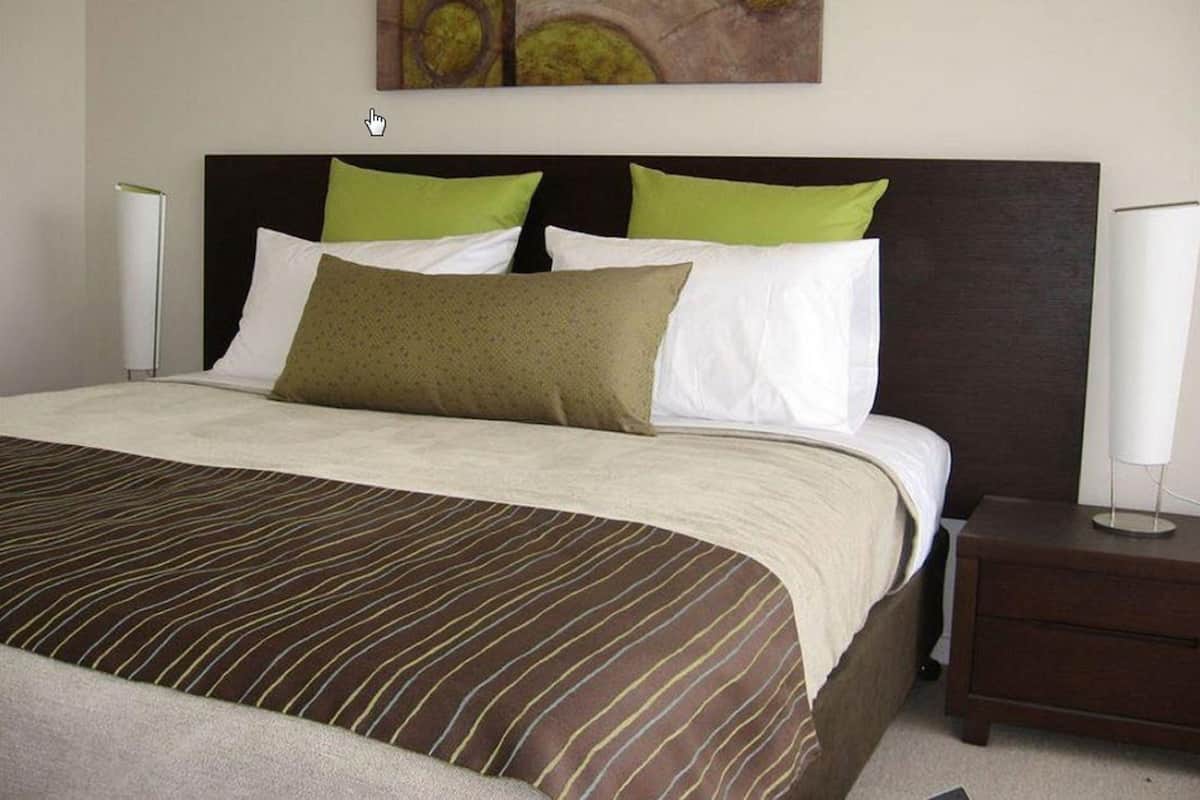 A hotel room with a large bed, green and white pillows, a dark headboard, and a painting on the wall.