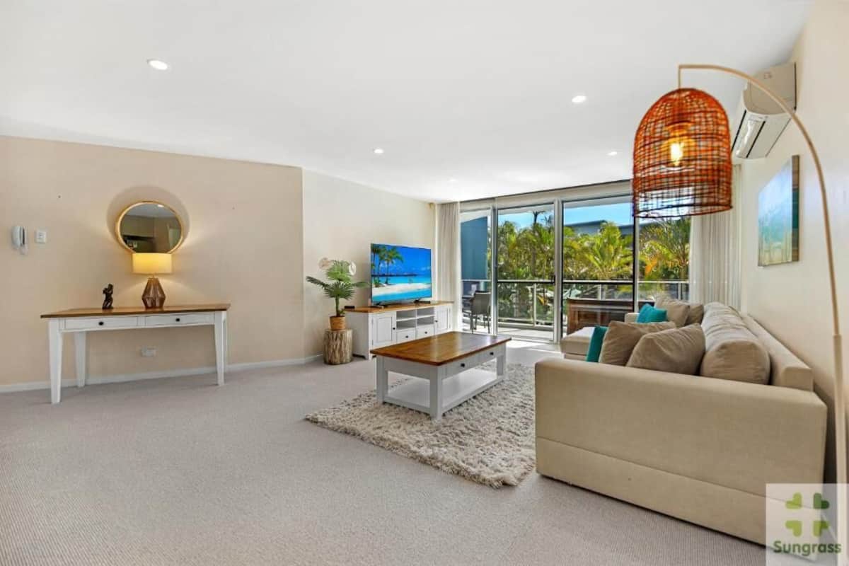 A modern living room with a sofa, a coffee table, a TV, and a large window with a view of palm trees.
