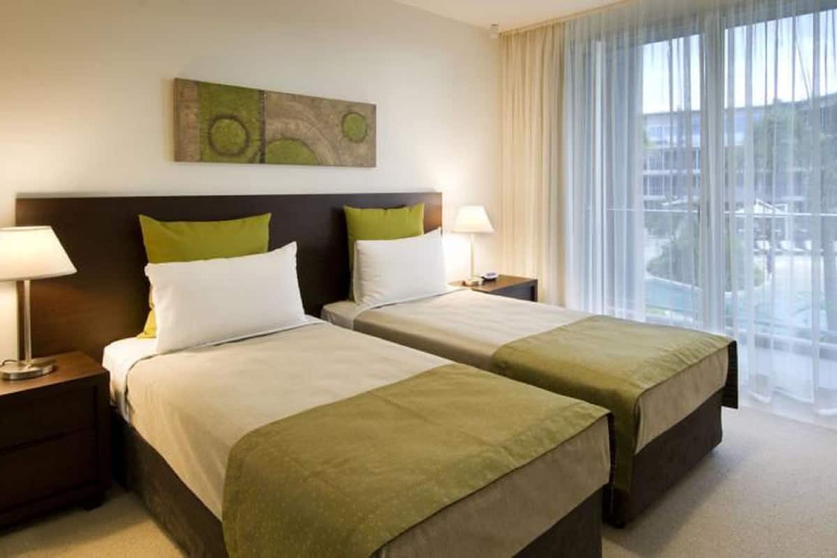 A hotel room with two beds, a nightstand with a lamp, a painting on the wall, and a large window with curtains.