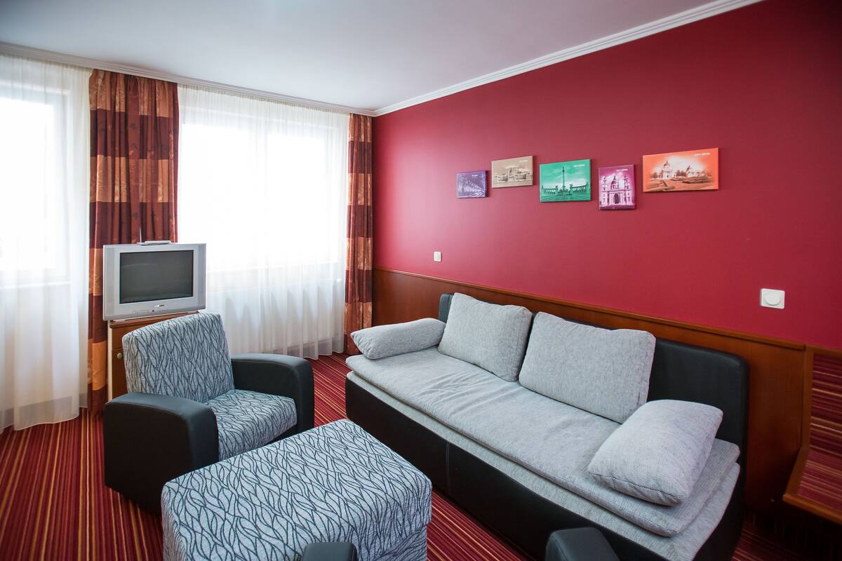 A hotel room with a red wall, a sofa, a chair, a TV, and a window with curtains.