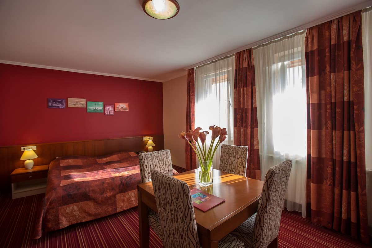 A hotel room with a bed, a dining table with chairs, a vase with flowers, and a wall with framed pictures.