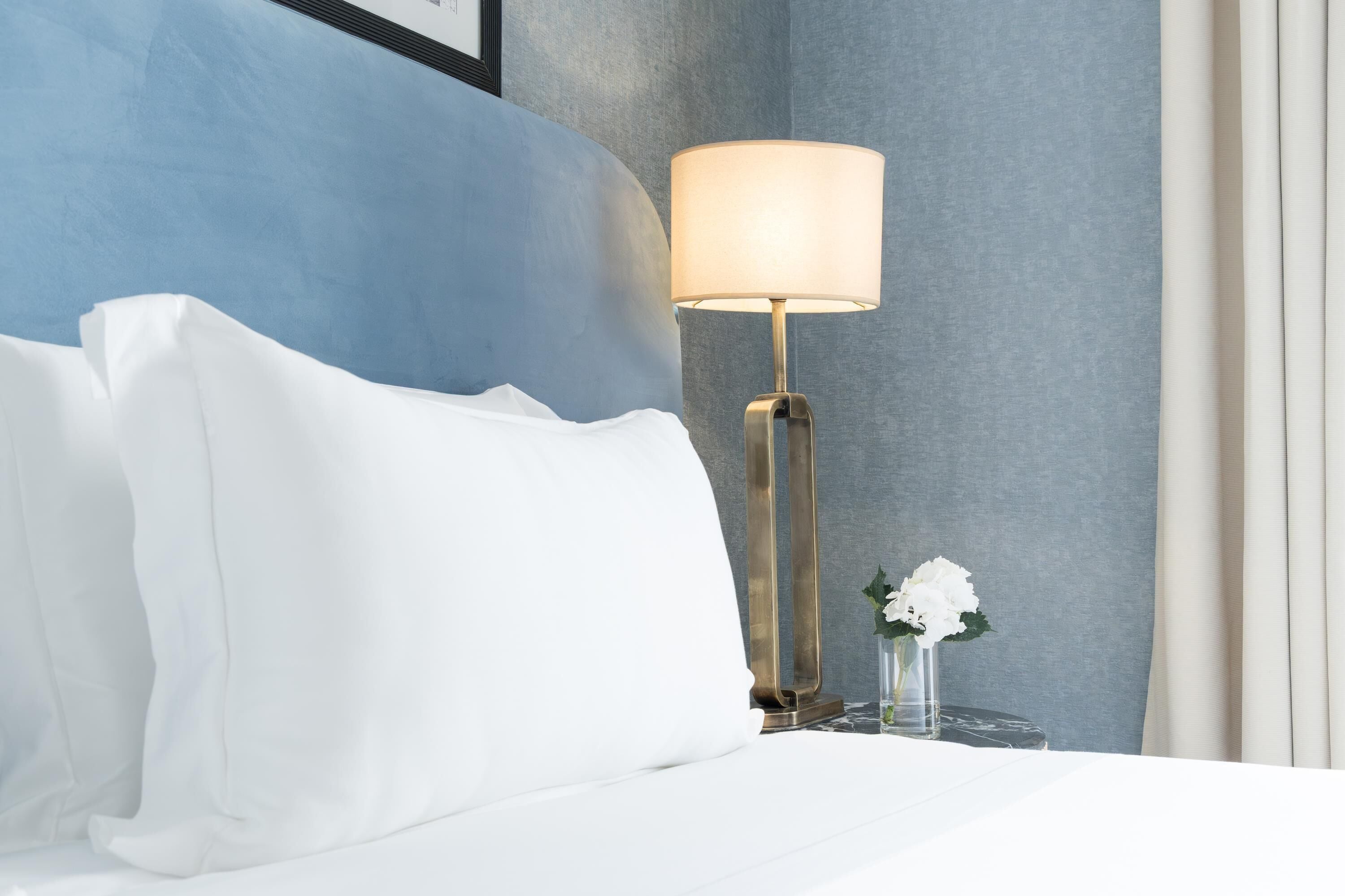 Una cama bien hecha con sábanas blancas, una lámpara de noche y un florero con flores blancas sobre una mesita de noche.