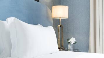 Una cama bien hecha con sábanas blancas, una lámpara de noche y un florero con flores blancas sobre una mesita de noche.