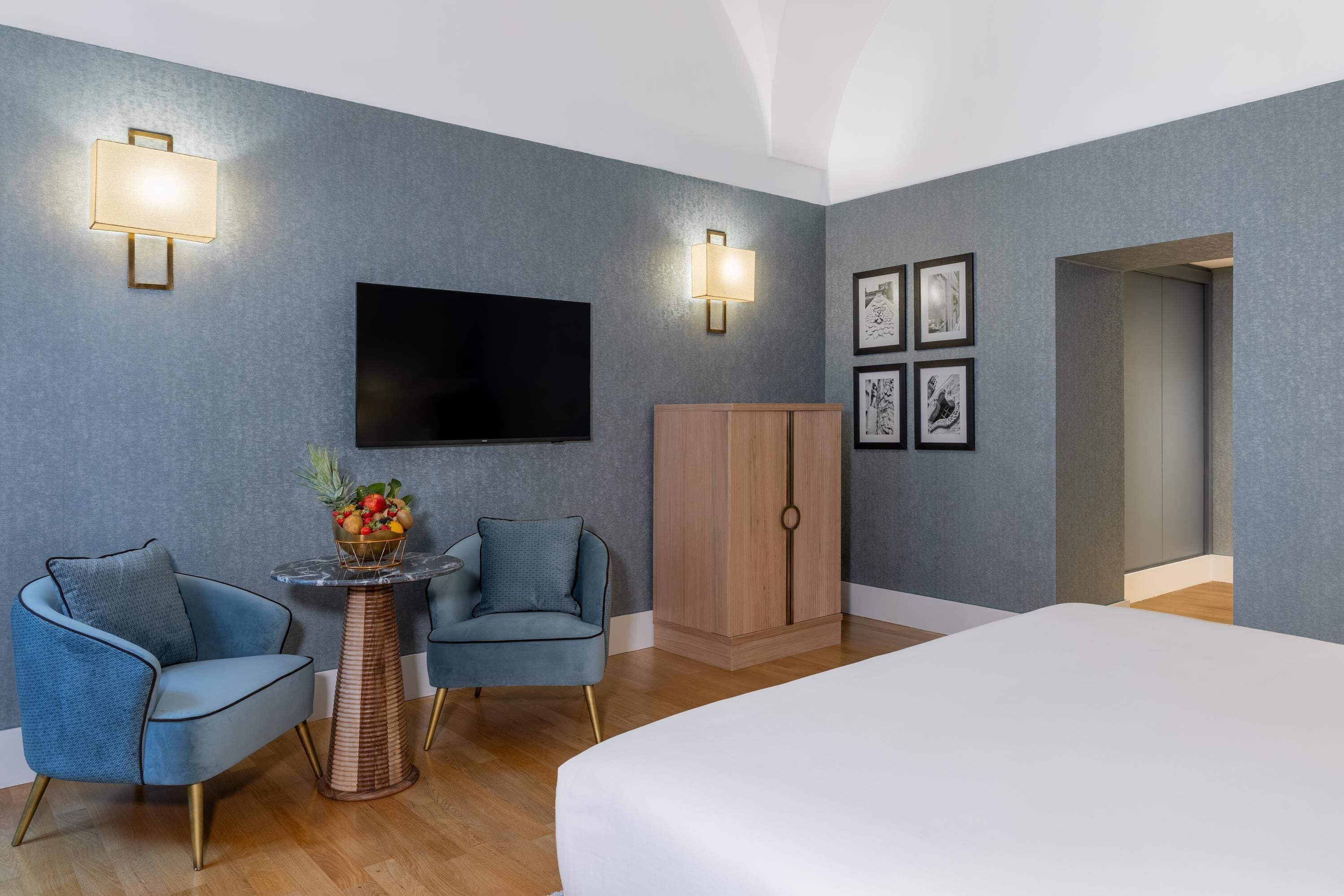 Ein modernes Hotelzimmer mit Flachbildfernseher, zwei Sesseln, einem kleinen runden Tisch mit Obstschale, einem Holzschrank und gerahmten Bildern an der Wand.