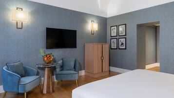 Ein modernes Hotelzimmer mit Flachbildfernseher, zwei Sesseln, einem kleinen runden Tisch mit Obstschale, einem Holzschrank und gerahmten Bildern an der Wand.