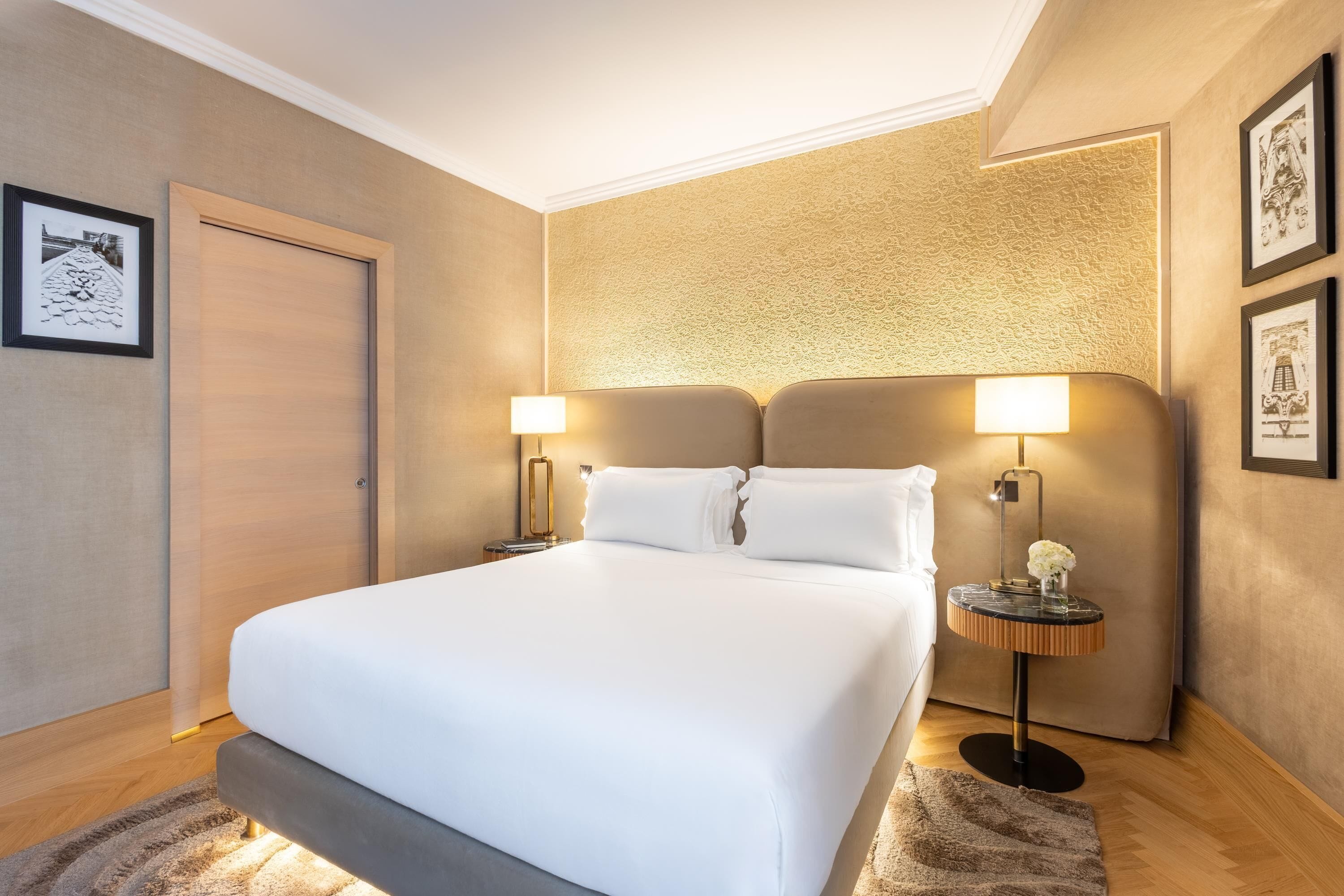 A hotel room with a large bed, two bedside tables with lamps, a door, and framed pictures on the walls.