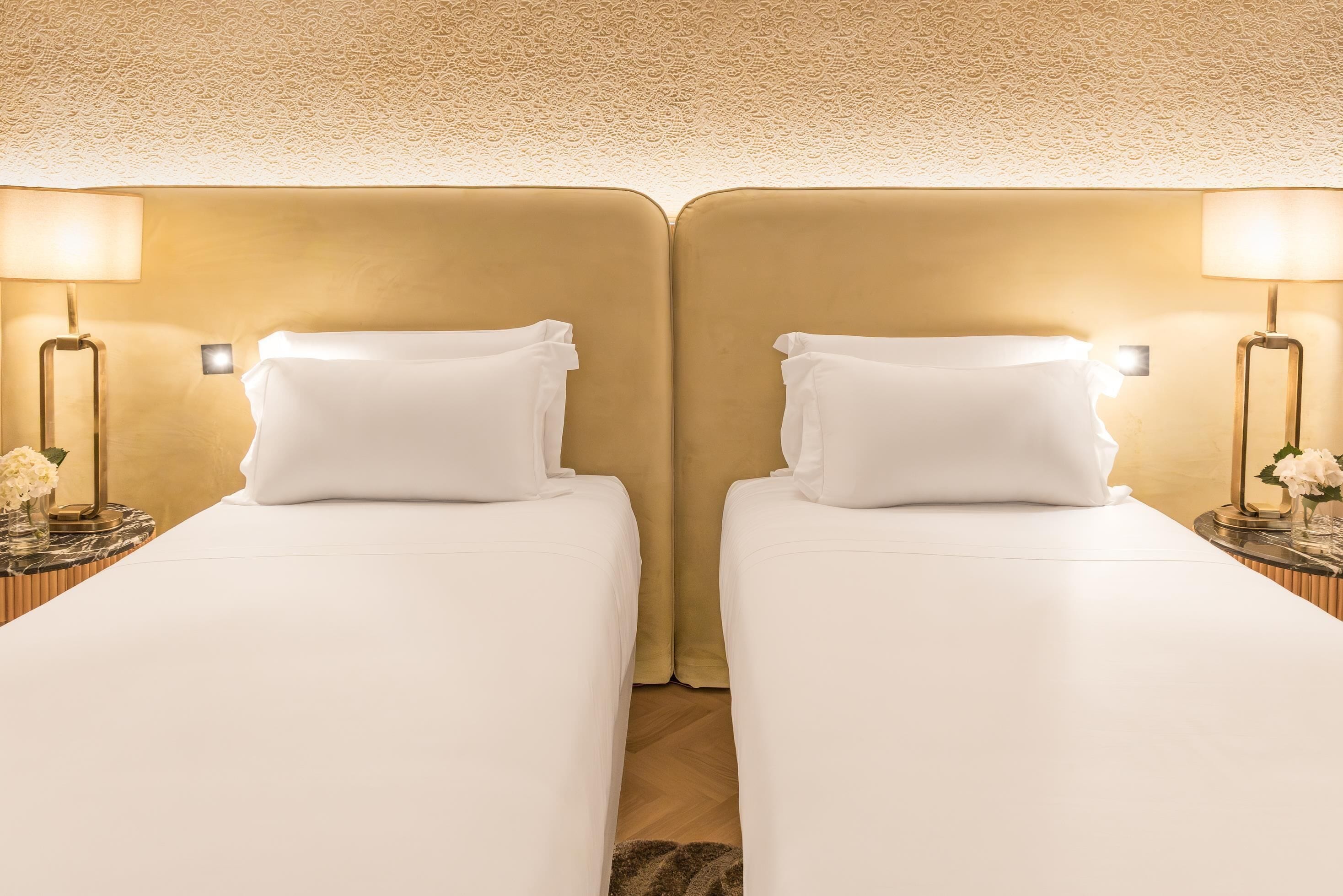 A hotel room with two beds, each with white linens and pillows, flanked by nightstands with lamps and small flower arrangements.
