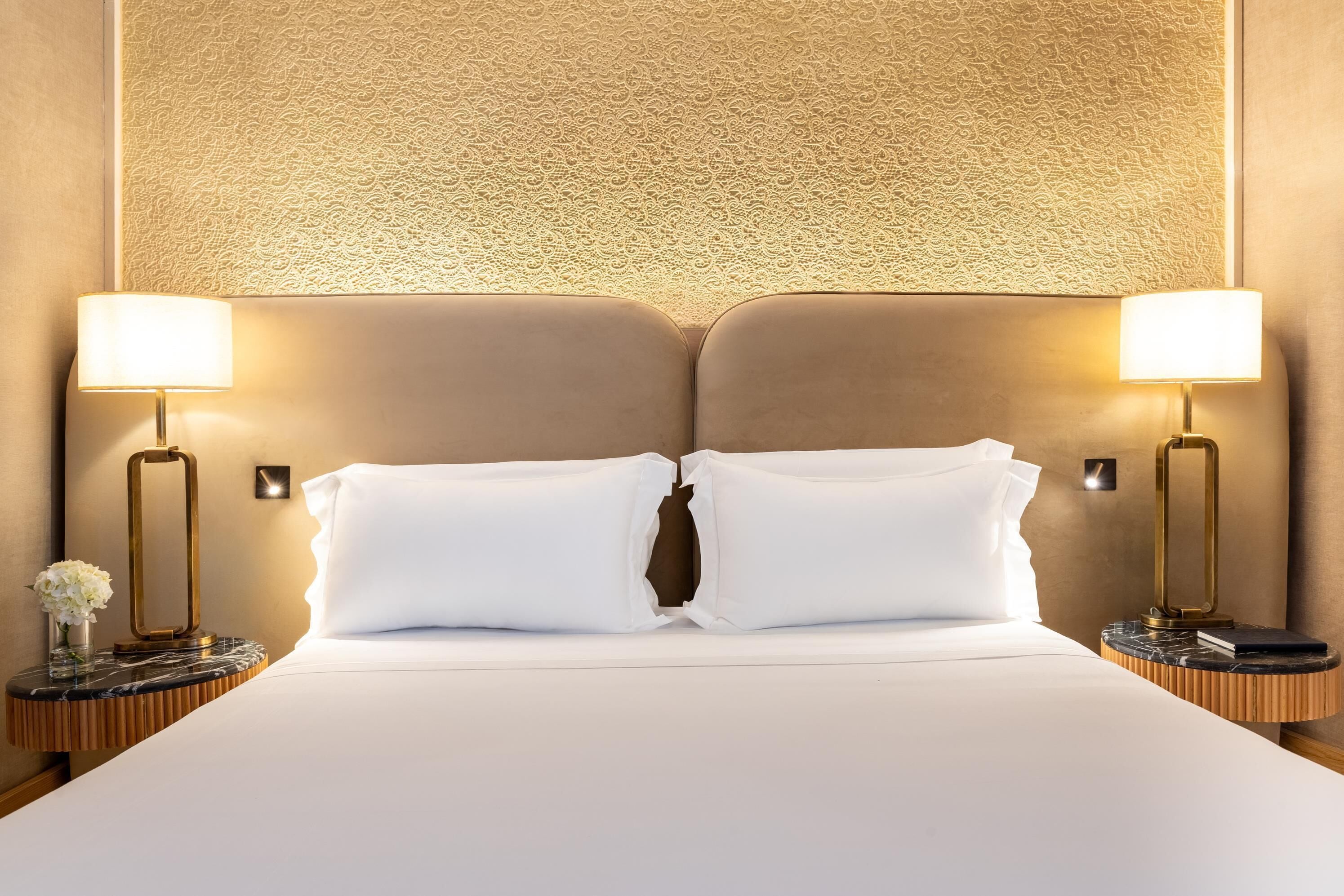 Una habitación de hotel con una cama grande, dos lámparas de noche, una mesita de noche con un florero y una pared texturizada detrás de la cama.