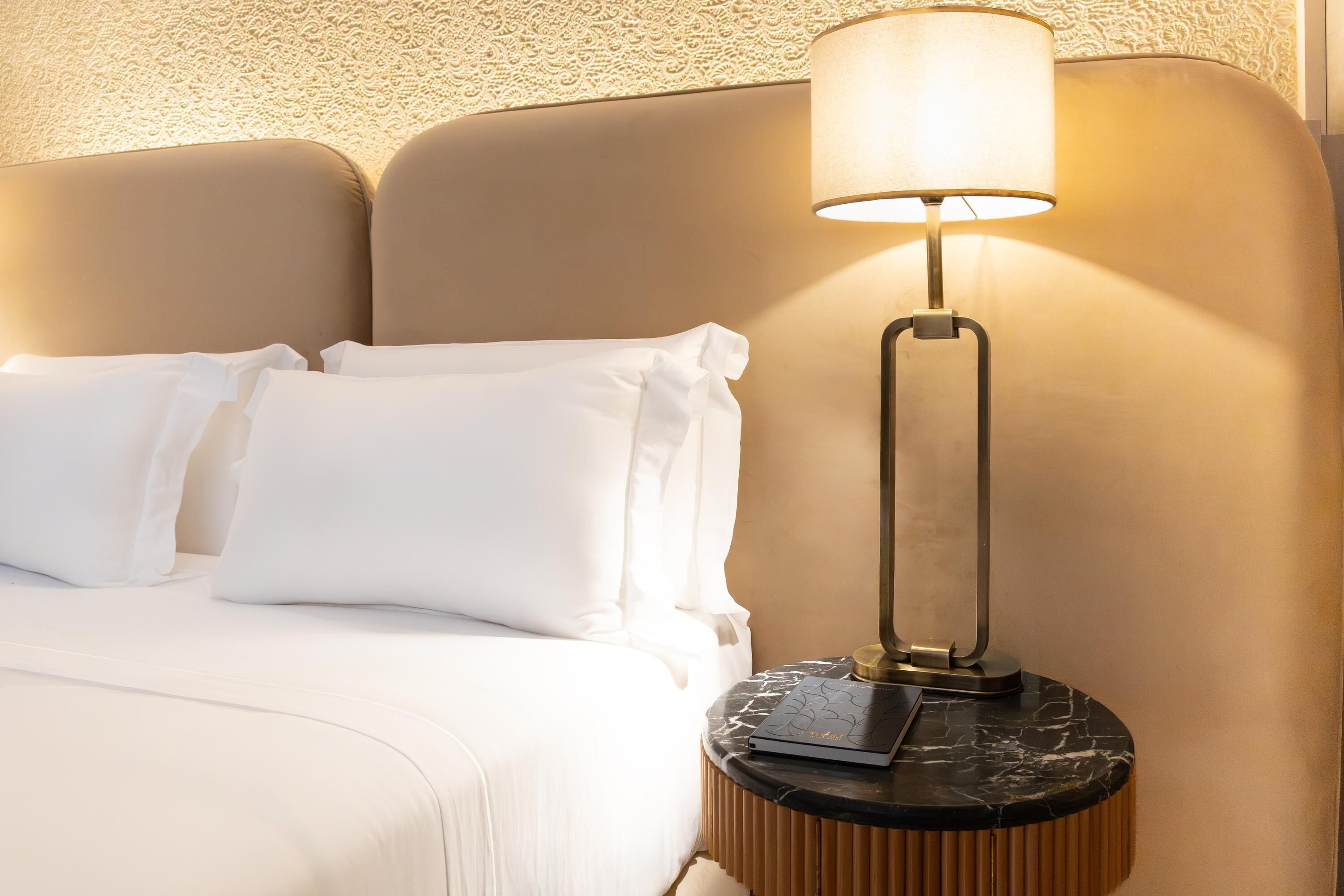 A bed with white linens, a round nightstand with a lamp, and a textured wall.