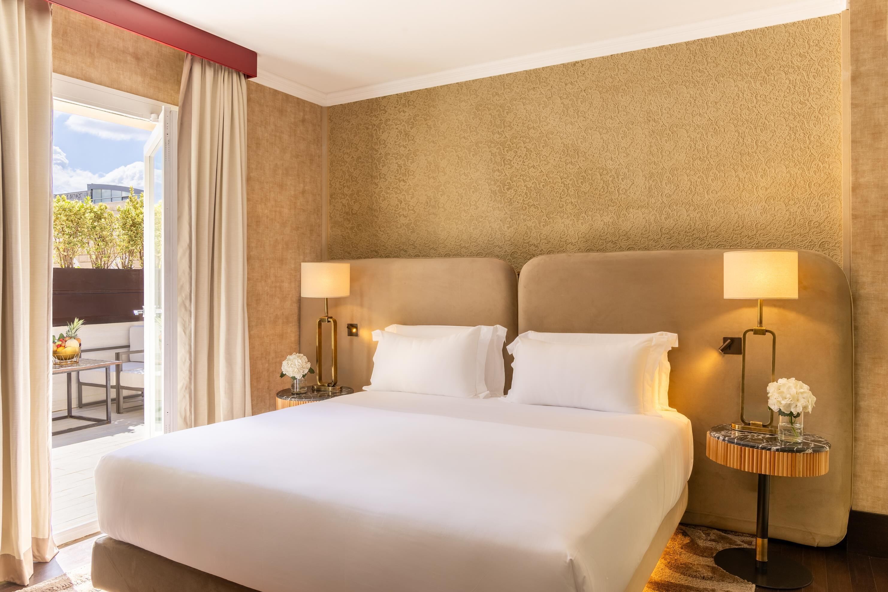 A hotel room with a large bed, two bedside lamps, a small round table with a vase, and a view of the outdoors.