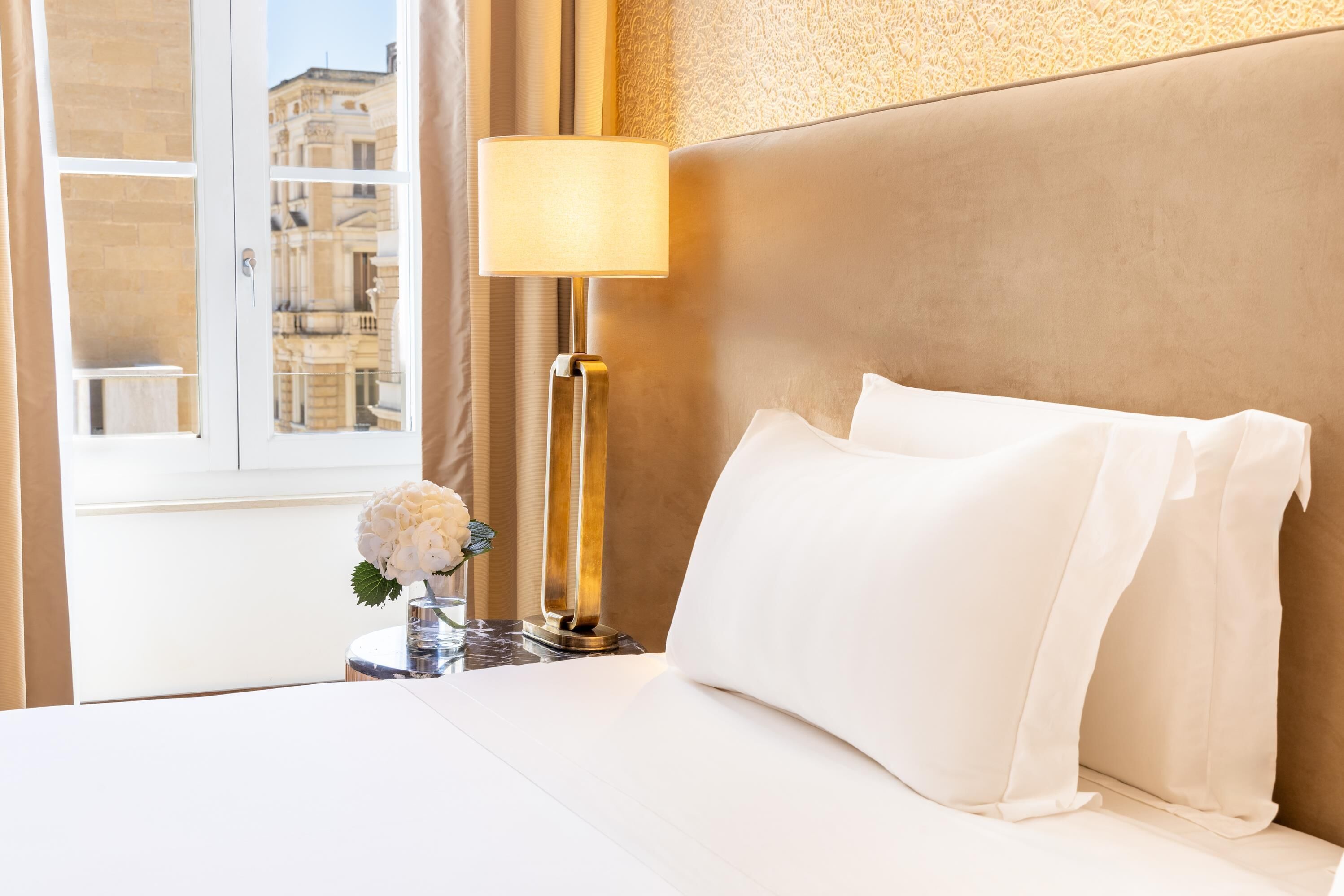 Habitación de hotel con cama, lámpara, un jarrón con flores y vistas a edificios a través de una ventana.