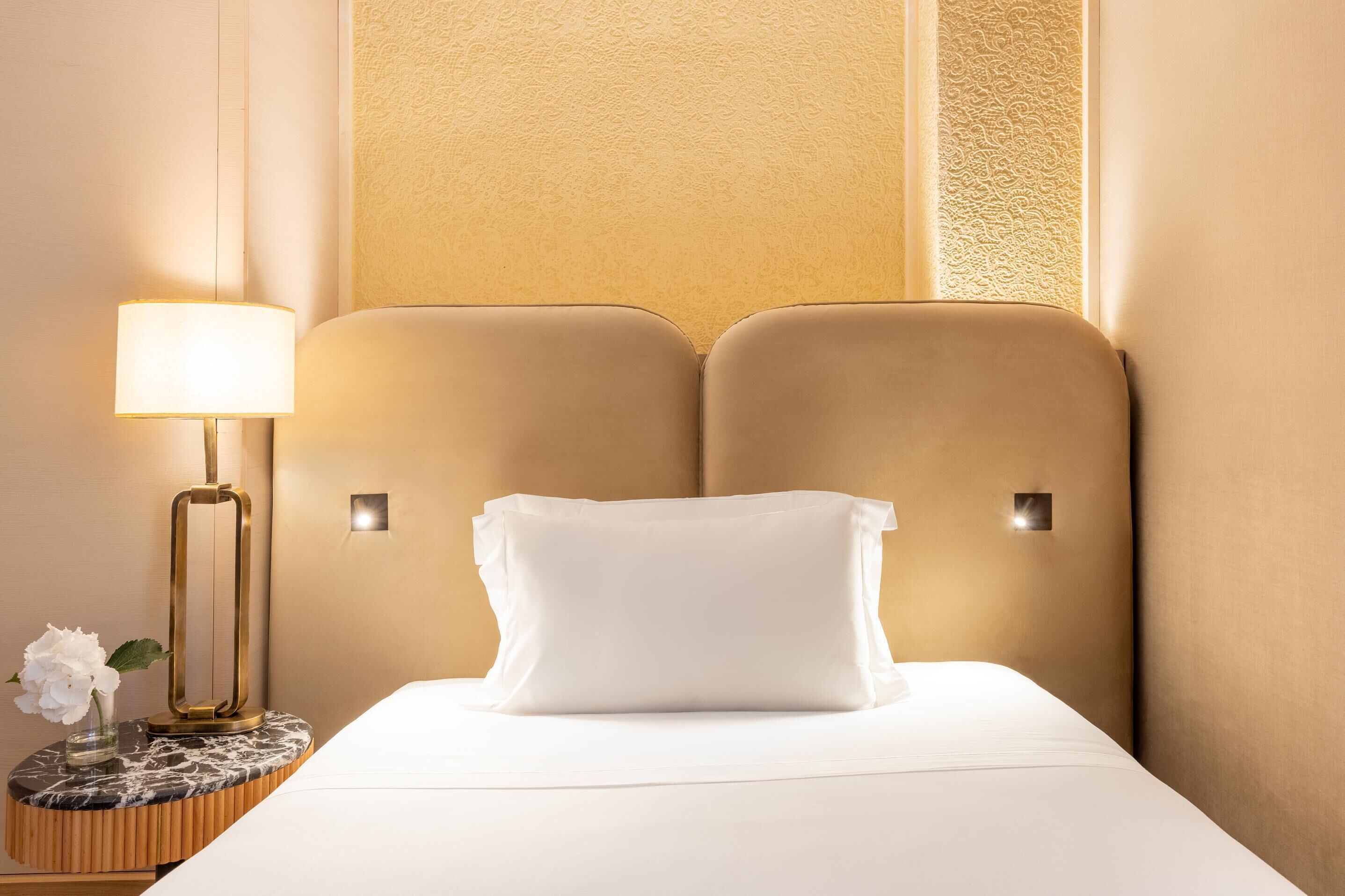 Habitación de hotel con cama, lámpara de noche, una mesita redonda con un florero y pared con textura.