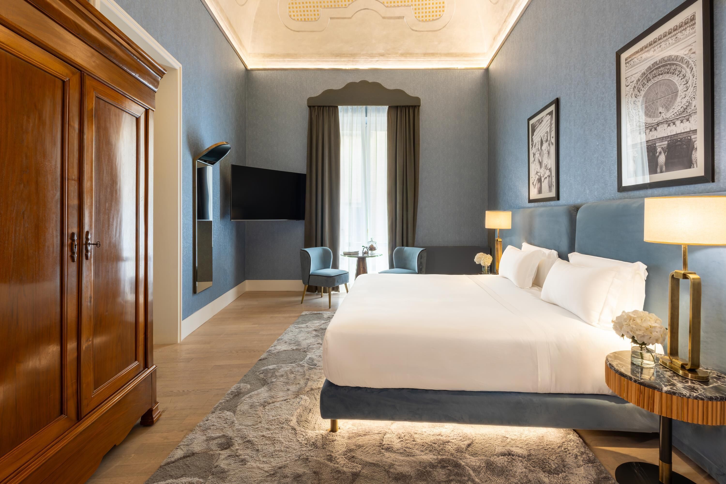 Habitación de hotel con una cama grande, un armario de madera, un televisor de pantalla plana, una mesita con una lámpara y una ventana grande con cortinas.