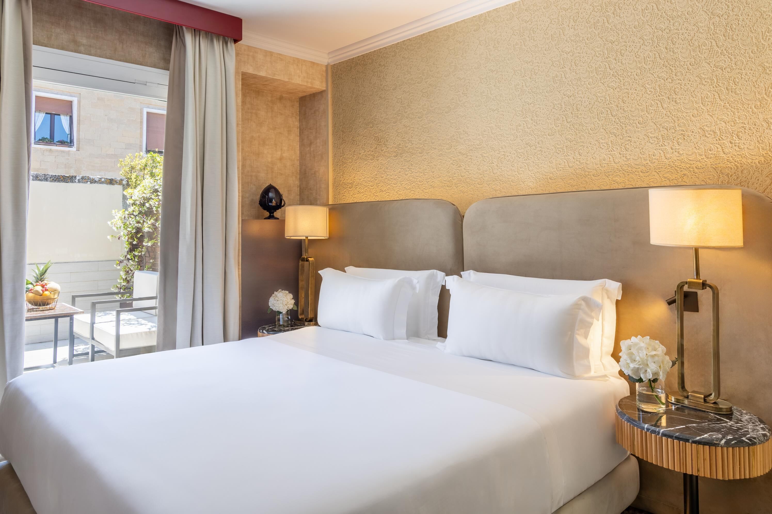 Een hotelkamer met een groot bed, twee bedlampen, een kleintje tafeltje met een bloevas, en uitzicht op het balkon.