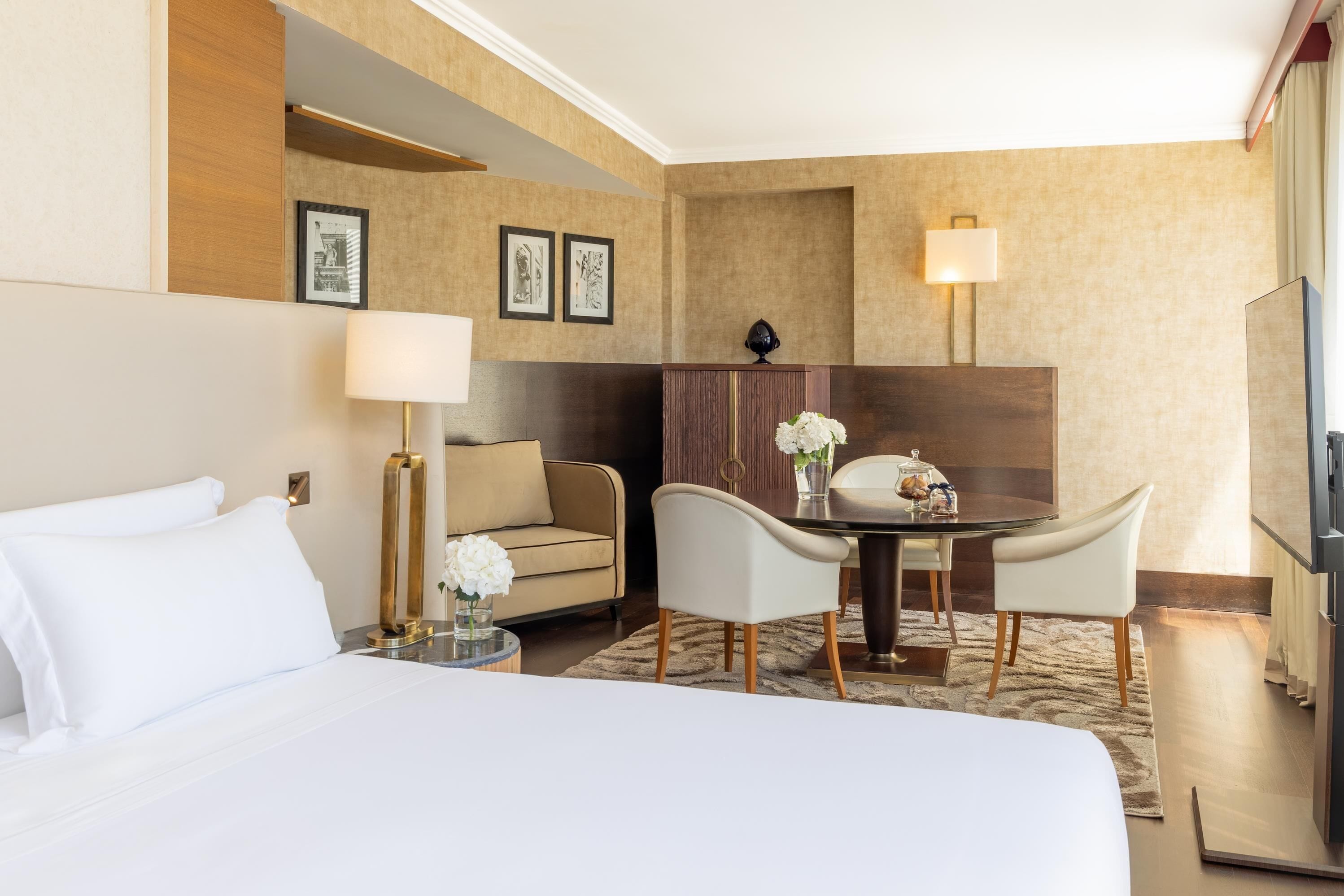A hotel room with a bed, a desk with chairs, a lamp, a television, and framed pictures on the wall.