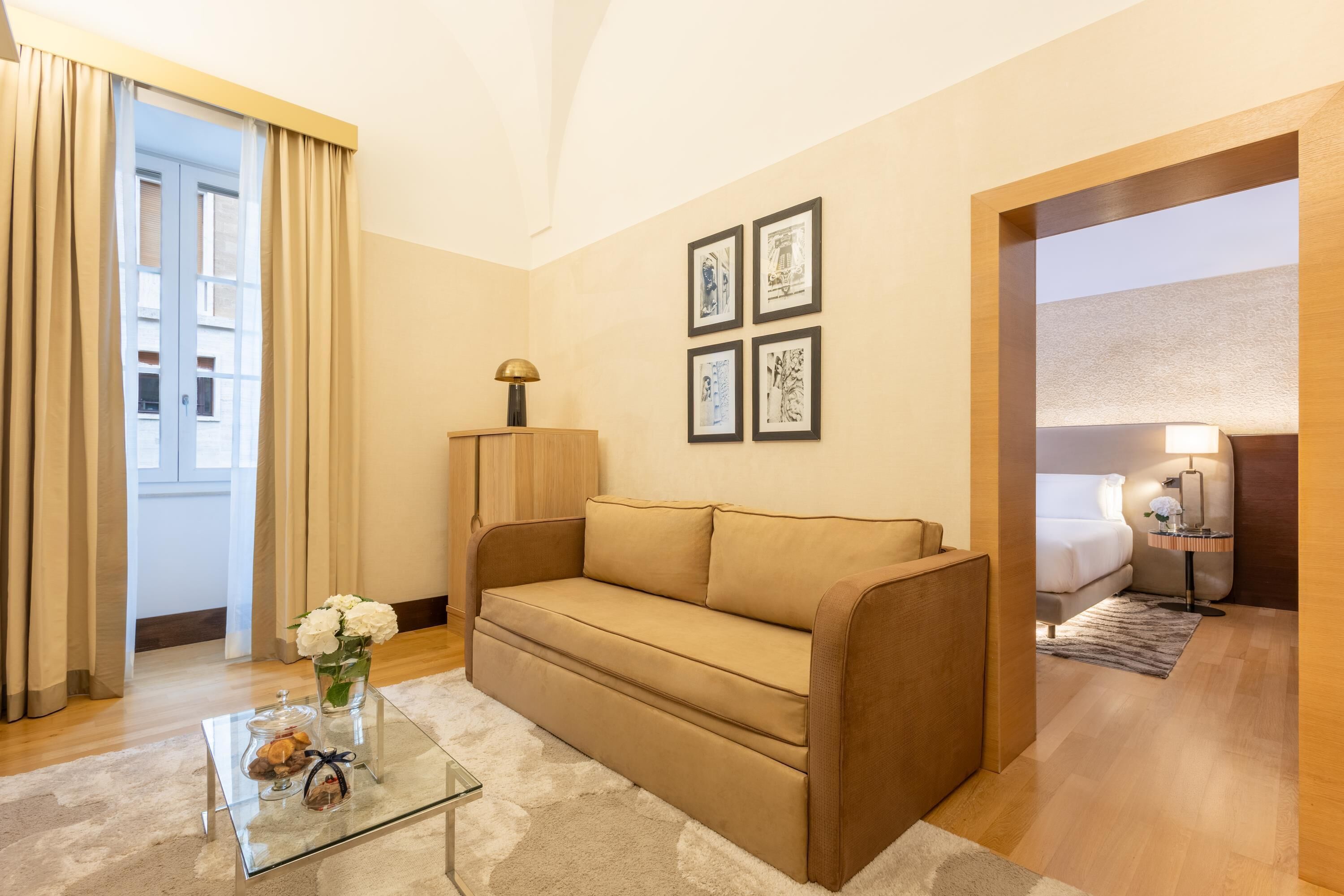Quarto de hotel com um sofá bege, uma mesa de centro de vidro com objetos decorativos, uma luminária e quadros nas paredes.